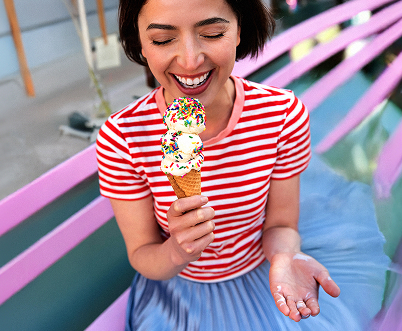 Eine junge Frau mit mittellangen, dunklen Haaren im rot-weiß-gestreiften Shirt lacht glücklich. In der Hand hält sie eine Eiswaffel mit drei Kugeln Vanille-Eis, die mit bunten Zuckerstreuseln bestreut sind.