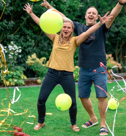 In einem Garten stehen ein Mann, eine Frau und ein Mädchen, alle in sommerlicher Kleidung. Sie werfen die Arme in die Luft und jubeln, um sie herum fliegen bunte Luftballons und Luftschlangen.