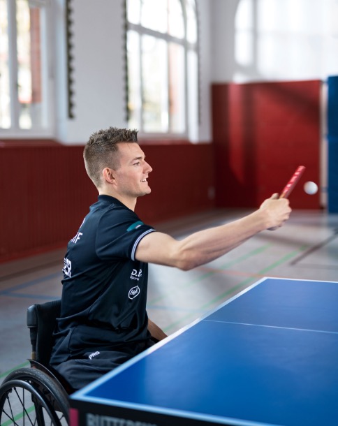 In einer Sporthalle spielt ein junger Mann Tischtennis. Er nutzt einen Rollstuhl, trägt dunkelblaue Sportkleidung und hat soeben einen Ball zurückgeschlagen.
