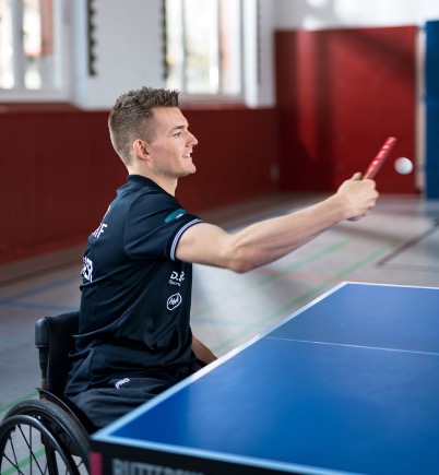 In einer Sporthalle spielt ein junger Mann Tischtennis. Er nutzt einen Rollstuhl, trägt dunkelblaue Sportkleidung und hat soeben einen Ball zurückgeschlagen.